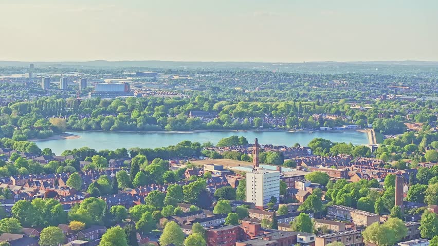 Drone ascend over Edgbaston Reservoir nestled among green spaces and residential structures during bright summer conditions in Birmingham, UK