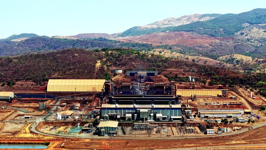 Nickel mining facility in Brazil, showing large industrial structures, processing plants, and surrounding mountain terrain under clear skies, aerial view