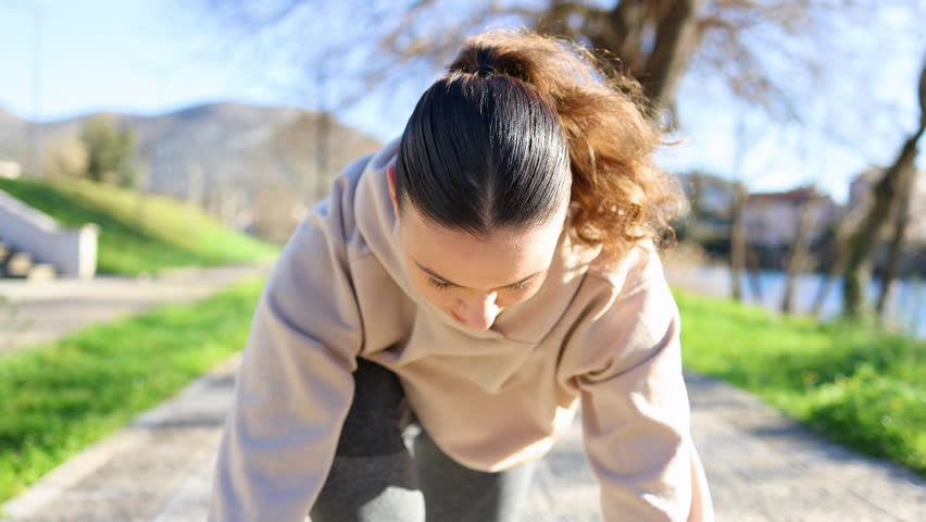 Energetic female runner crouched in a starting block position, gazing up and flashing a smile towards the camera. Young athletic woman gearing up for a sprint in an outdoor setting