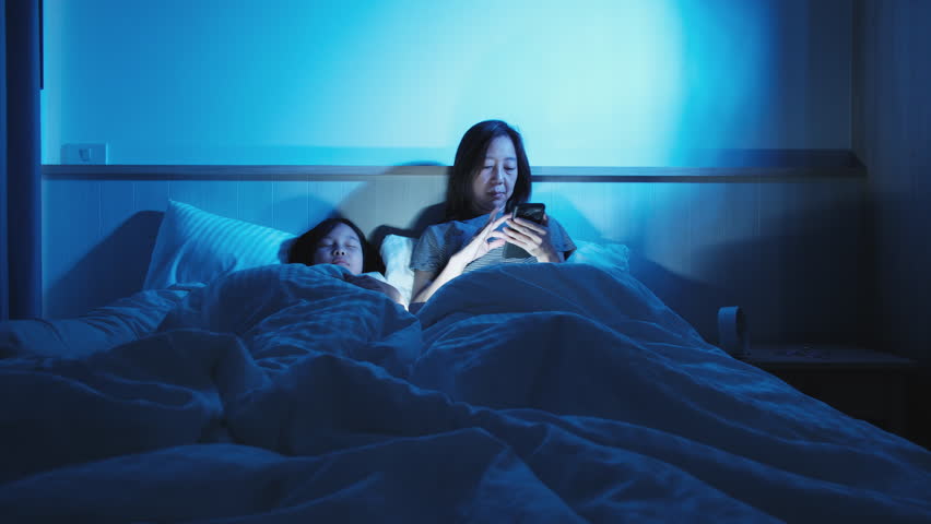 Asian single mum is sleepless, using mobile phone before sleep, social media addiction, insomnia all night near a daughter in bedroom, mature and menopausal woman, widow middle-aged mother lifestyle.