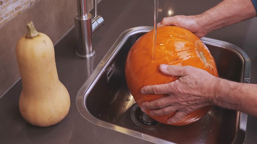 Man rinses bright orange pumpkin under running water in kitchen sink, preparing for seasonal cooking.