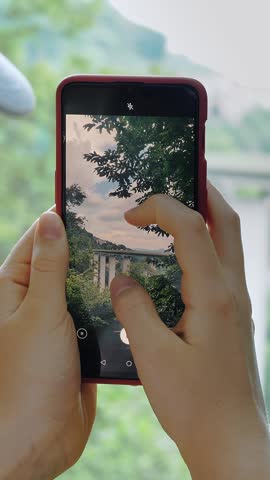 Close-up. A girl in the park during summer, capturing a beautiful landscape on her mobile phone. A bridge for cars and the Alps in the background.