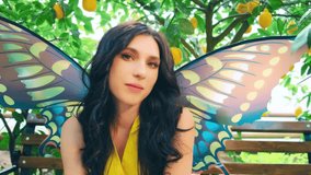 Fabulous fantasy girl elf, yellow dress, butterfly art wings. Portrait fantasy happy woman smiling face fairy long loose wavy dark hair, sitting on wooden bench. Backdrop garden, lemon trees green. - Powered by Shutterstock - Get 15% off with code: PIKWIZARD15