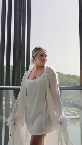 Bride posing in Hotel Room. Smiling Schuman Sharing Luhury Accessoris and Vgite Silk Bathrobe with Featgers. Preparation for Wedding Ceremony during Morning Time