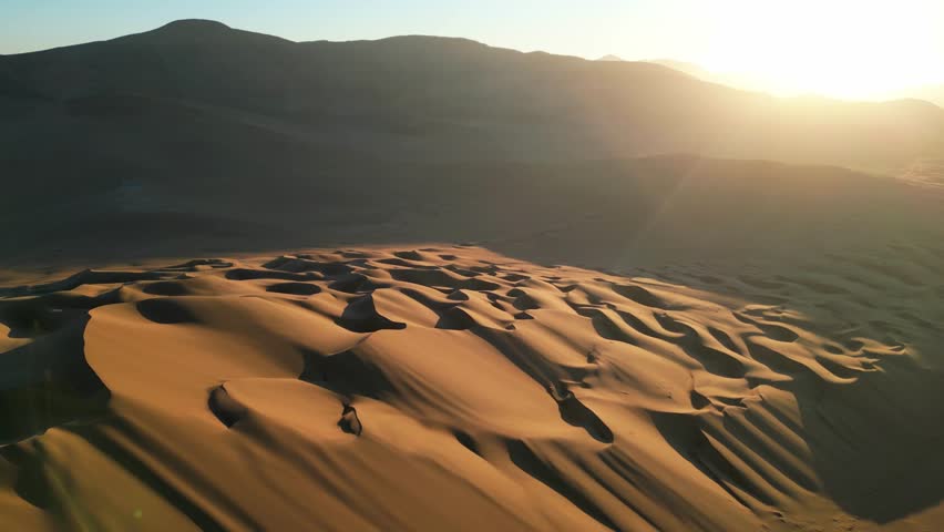 Aerial drone view of golden sand dunes in the southern Atacama Desert, glowing in warm sunset light.