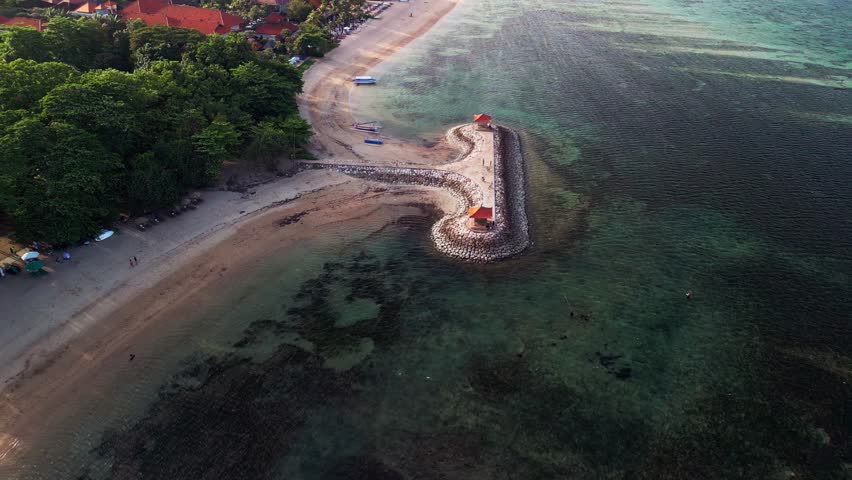eautiful Sanur Beach in Bali seen from above with wide sandy coast shimmering blue water protective coastal barriers and red roof gazebos on top filmed in a tropical drone video style.