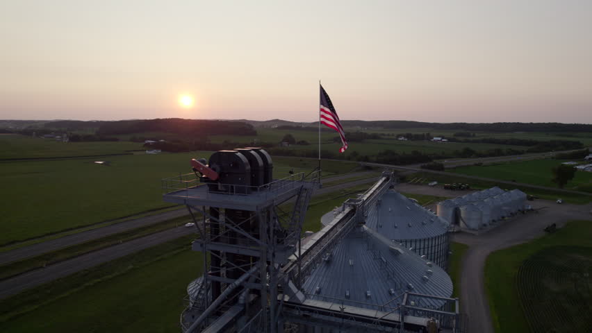 Drone footage of a rural grain storage facility with large metal silos and an American flag at sunset over surrounding farmland.