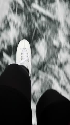 Top view of female legs in white figure skates are skating on thick ice on lake, close up. Woman in skates is gliding on frozen river. Vertical video