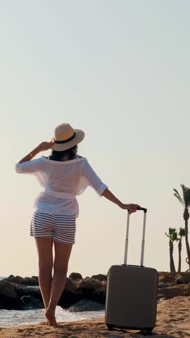 Back view of young woman in summer clothes and sun hat, holding travel bag, looking at the sea, enjoying the sunrise, against the background of the beach and palm trees.