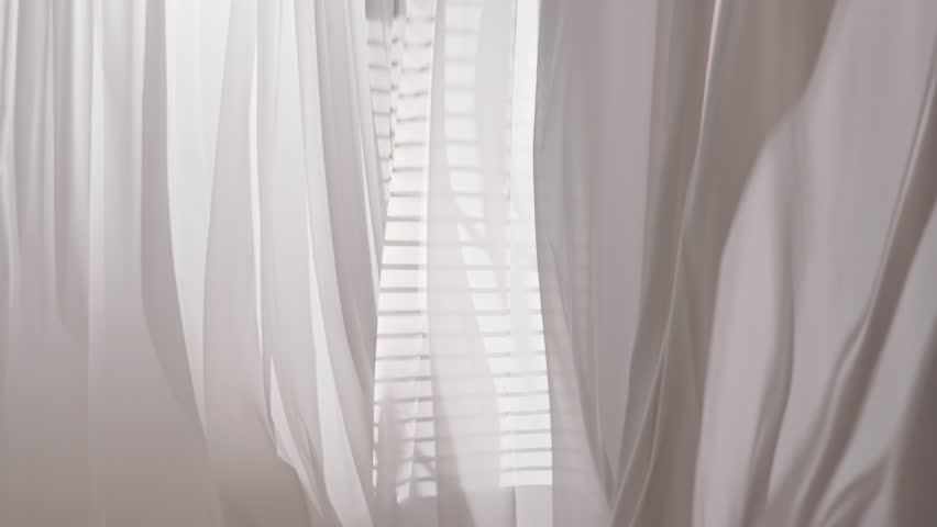 Transparent silk curtains in daylight effect floating from an open window, shadows from window blinds reflected on the textile.