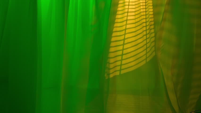 Transparent silk curtains in green neon light floating from an open window, yellow shadows from blinds reflected on the floor and textile.