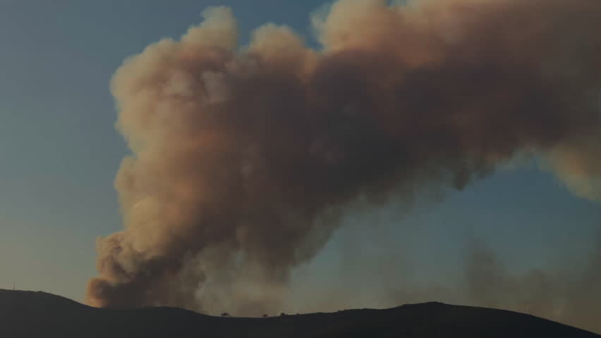 A massive wildfire burns on a mountain, sending huge plumes of smoke high into the sky, visible from a distance.