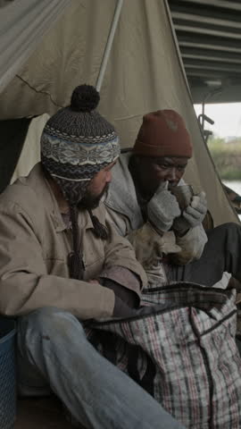 Vertical view of Caucasian homeless man taking aluminum can out of plaid bag and downing last bit of beer, Black beggar with metal cup watching him outdoors