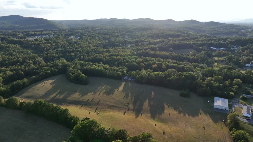 Hay bales on grass field of American countryside farm at sunset. Aerial wide shot. Peaceful nature landscape with mountains in the evening. Virginia, USA. Summertime with house and barn.