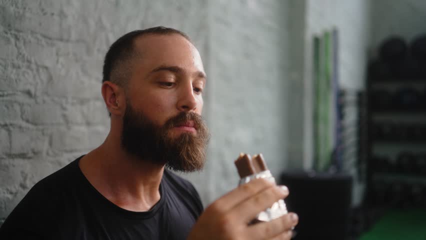 Mid adult bearded man in black t-shirt examining chocolate protein bar during fitness recovery moment in modern training facility 