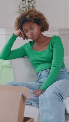 Young African American woman sits on sofa surrounded by scattered items, visibly upset and disappointed after unboxing her recent online shopping order that did not meet her expectations.