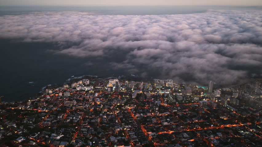 A stunning aerial view of Cape Town at dusk, where glowing city lights meet the vast ocean, as a dramatic blanket of clouds rolls in from the horizon.