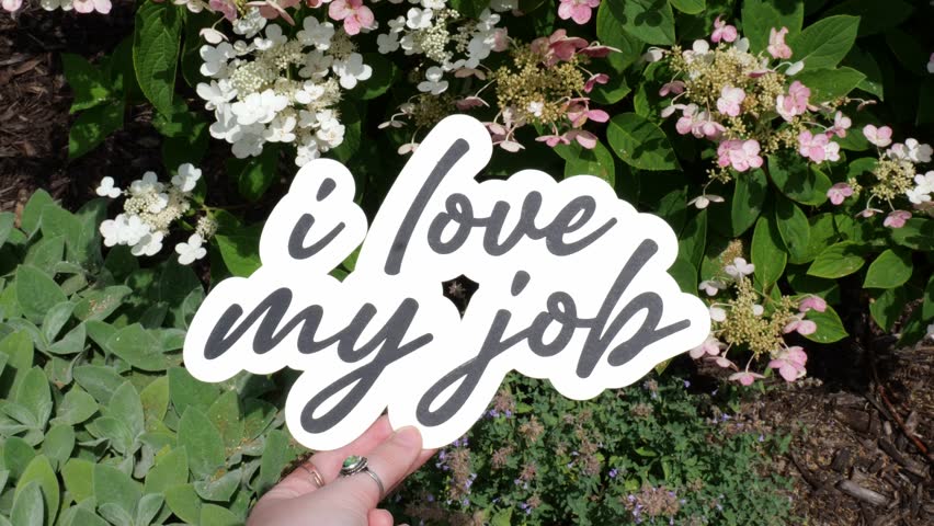 Young Woman Holds Up message of I Love My Job Wording in front of White and Pink Flowers in a garden in Summer 4K Video Stock Footage