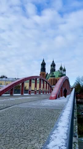 Priest in a long black robe walking quickly across the famous red bridge in Poznan, Poland, heading toward the historic Cathedral Island (Ostrów Tumski) with church towers in the background on a sunny