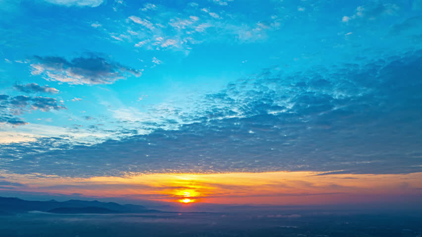 Aerial shot of a gorgeous sunrise. A panoramic view of dramatic mountain peaks silhouetted against a vibrant sunset sky, with fiery hues of red, orange, and pink blending into the fading blue. 