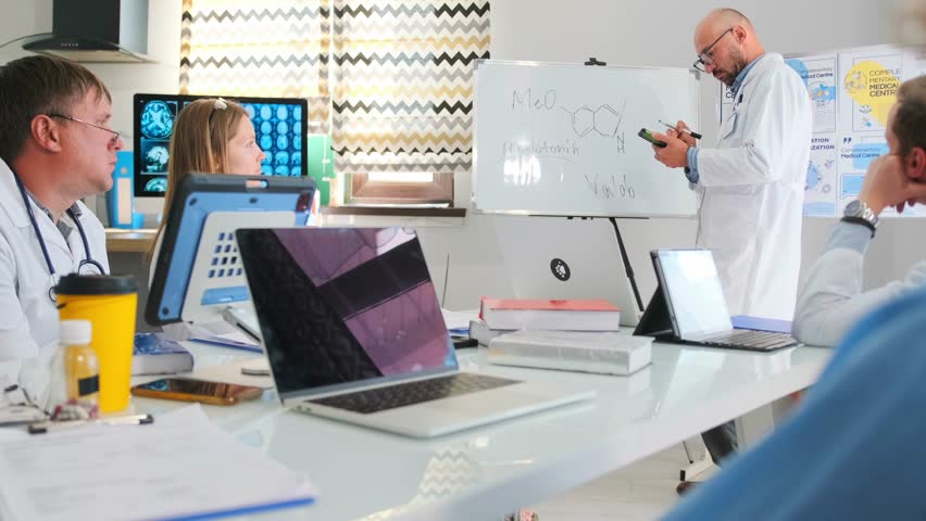 Bald pharmaceutical representative in white coat writes with marker on whiteboard holding phone during hospital seminar. Medical staff listen to lecture about new drug and healthcare innovations.