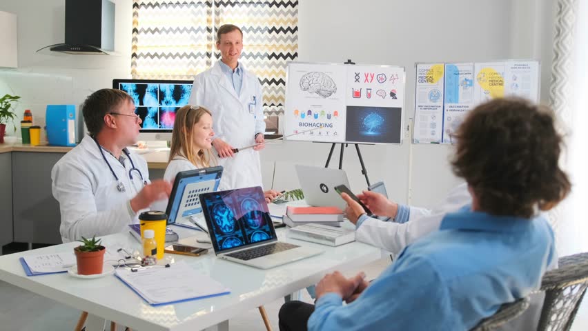 Doctors and interns participate in a hospital seminar. Lecturer uses a pointer to highlight brain diagrams and charts about biohacking. Participants watch during the educational session. 