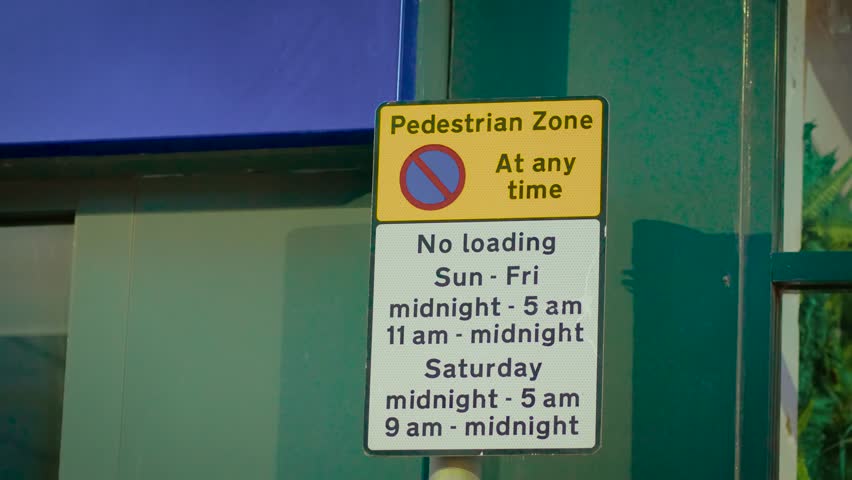 Sign indicating no parking in pedestrian zone mounted on wall near storefront. Board displaying restriction with text pedestrian zone at any time no loading. Panel informing drivers about limited