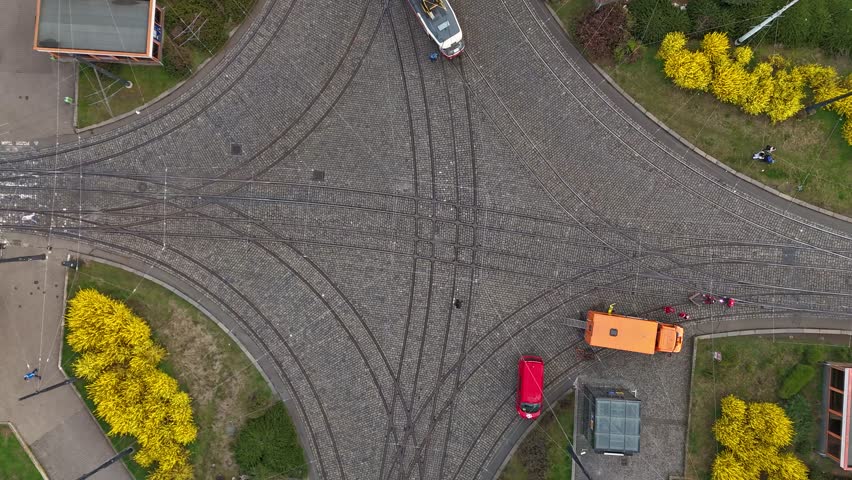Aerial drone view of an urban intersection with tram tracks, moving traffic and pedestrians, showcasing mobility, public transport network, road infrastructure, and dynamic street life.