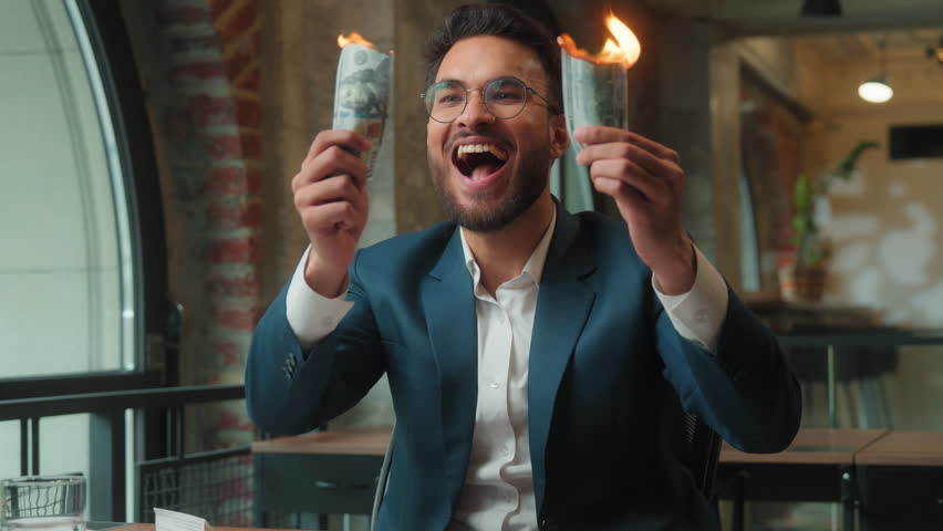 Happy Hindu business man burning money in office laughing male Arabian Indian businessman crazy mad executive entrepreneur CEO investor fooling having fun burn cash dollars salary spending finance