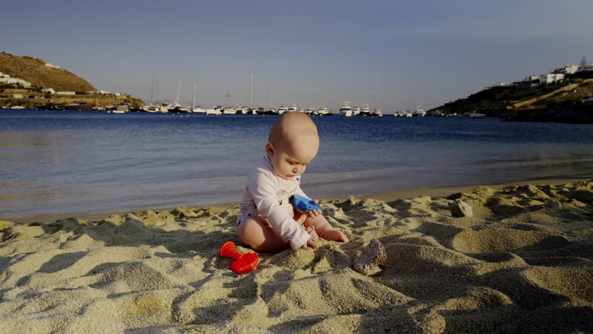 Young boy playing with colorful toys on sandy beach, surrounded by gentle waves and distant boats, enjoying a sunny day by the ocean with joyful expression