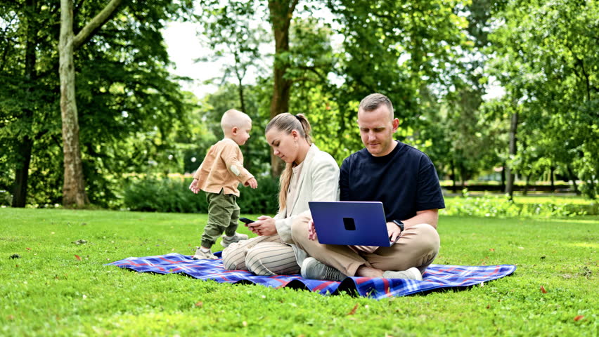 A young family couple works outdoors with a laptop while their child plays nearby. A happy family couple with a child are relaxing outdoors and working remotely on a laptop.