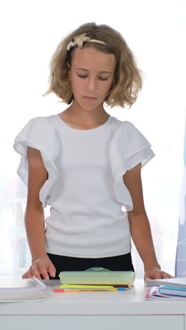 Vertical video. Young girl neatly placing pens pencils and supplies into a pink schoolbag on a white desk in her study room