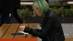 Woman with green hair writes notes in a notebook while using a calculator on her smartphone, sitting outdoors at a wooden table in the city. Side view portrait. Horizontal 4k footage - Powered by Shutterstock - Get 15% off with code: PIKWIZARD15