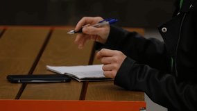 Woman hands writes notes in a notebook while using a calculator on her smartphone, sitting outdoors at a wooden table in the city. Close up side view. Horizontal 4k footage - Powered by Shutterstock - Get 15% off with code: PIKWIZARD15