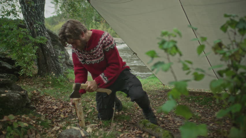 Survivalist chopping wood with hatchet while preparing to make fire near tarp shelter on riverbank in forest