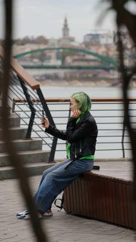 A young woman with green hair in a leather jacket and jeans sits on the embankment, holding a smartphone and taking a selfie with the city and bridge in the background. Vertical 4k footage