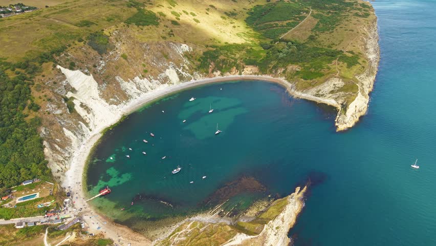 Drone footage orbiting Lulworth Cove, revealing boats on turquoise water in summer
