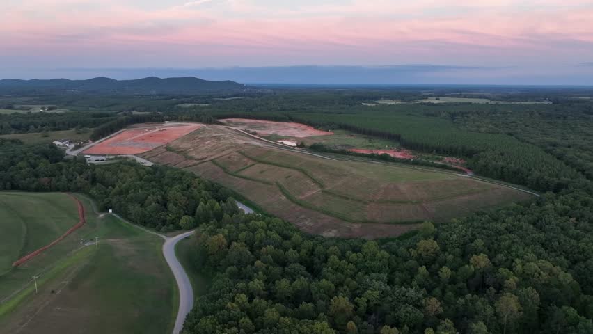 Aerial wide shot of an open landscape in America with red soil excavation, farmland, forest, winding road and distant mountains under soft pastel sunset sky. Peaceful scene in USA