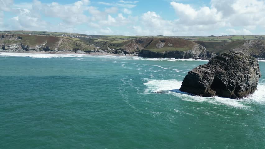 Aerial view of rugged coastline at Trebarwith Strand, Cornwall. The turquoise water meets steep cliffs with waves crashing around a large rock formation under a bright, clear sky.