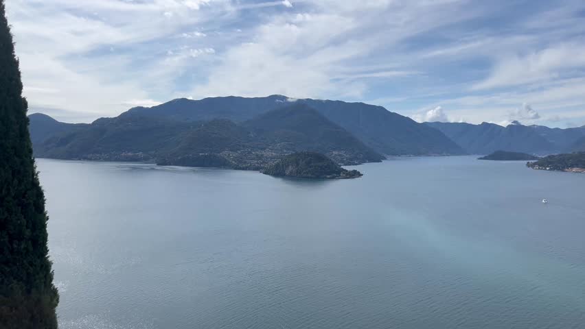 Panoramic view of the picturesque Lake Como in Italy, with its calm waters reflecting a clear sky with light clouds. The majestic mountains of the Alps frame the horizon, creating an exciting backgrou