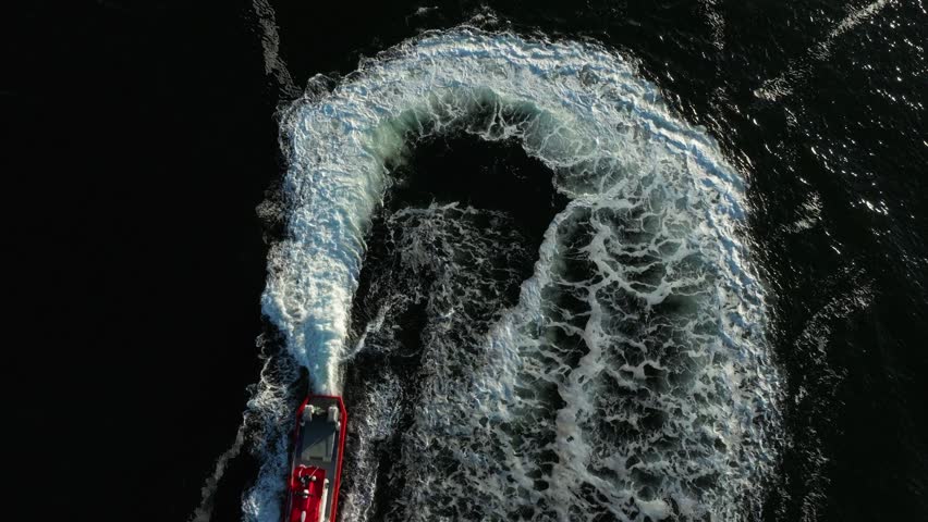 A red boat swiftly maneuvers through water, leaving behind a spiral pattern of white foam and waves under a clear blue sky. The scene captures the thrill of boating.