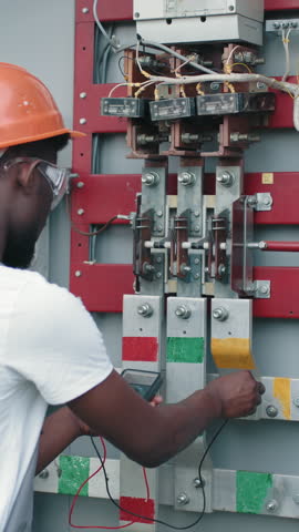 An electrician in a hard hat and safety glasses uses a multimeter to inspect an electrical panel, ensuring safety and functionality.