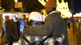 Multiethnic couple embracing while walking through a festive christmas market at night, surrounded by twinkling lights - Powered by Shutterstock - Get 15% off with code: PIKWIZARD15