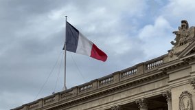 A video shows the flag of France fluttering gently in the wind atop a majestic, classical building. The blue, white, and red vertical stripes are clearly visible as the flag billows against a dramatic - Powered by Shutterstock - Get 15% off with code: PIKWIZARD15