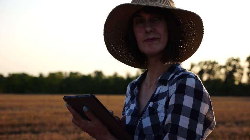 Female agronomist in straw hat monitoring harvest on barley field at sunset. Adult farmer using digital tablet on wheat meadow at dusk. Beautiful scenic landscape. Concept of agricultural business
