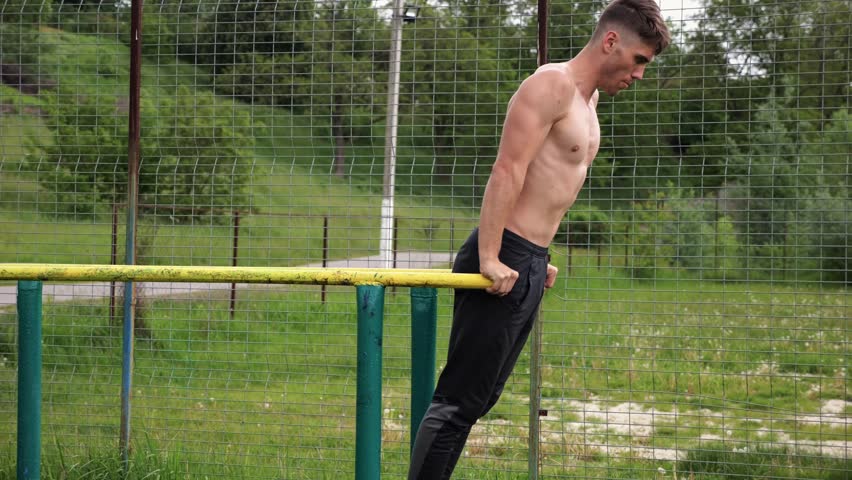 Young sportsman doing push ups on parallel bars at nature. Strong sporty man exercising at sports ground. Male shirtless athlete training hard outdoor. Concept of active sportive lifestyle. Slow mo