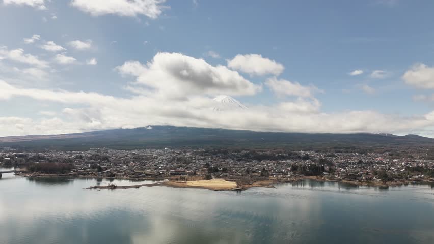 Aerial view of Fujikawaguchiko in Fuji, Japan, with Mount Fuji rising above the clouds, a calm lake in the foreground, and a densely built town spreading across the landscape under a partly cloudy sky