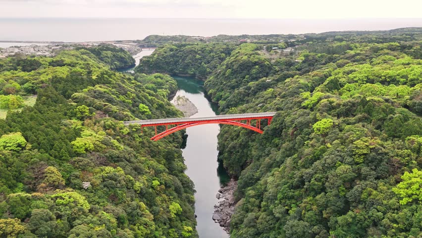 Matsumine Bridge spanning river gorge with red structure contrasting verdant forest landscape in Yakushima, UNESCO World Heritage Site capturing natural infrastructure integration, drone push in