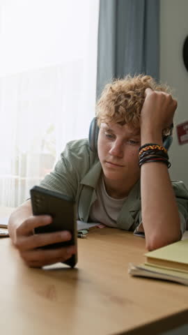 Vertical shot of Caucasian young man holding smartphone and mindlessly watching videos online while sitting at desk distracted from studying
