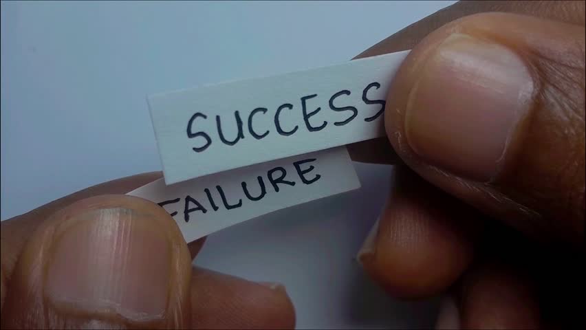 A pair of hands holds a paper with the word "FAILURE." As the paper is lifted, another paper beneath it reveals the word "SUCCESS." The hands place the "SUCCESS" sign in the center of the frame.